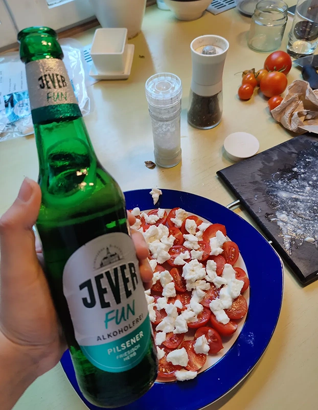 Eine Jever Fun Flasche wird vor einem Teller mit Tomaten und Mozzarella gehalten. Im Hintergrund liegen Küchenutensilien.