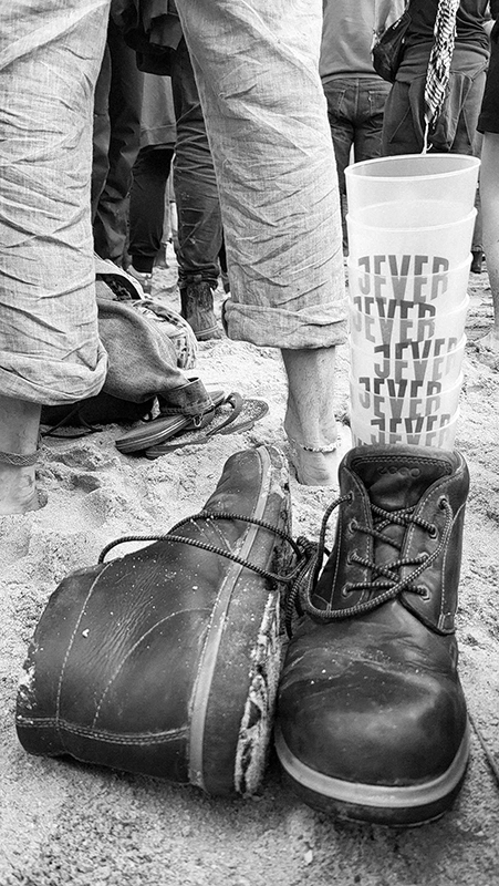 Ein Paar Stiefel steht im Sand neben gestapelten Jever Trinkbechern. Im Hintergrund stehen barfüßige Menschen.