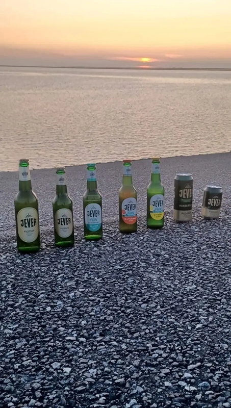 Mehrere Jever Bierflaschen und Dosen stehen nebeneinander auf Asphalt. Im Hintergrund ist das Meer bei Sonnenuntergang zu sehen.