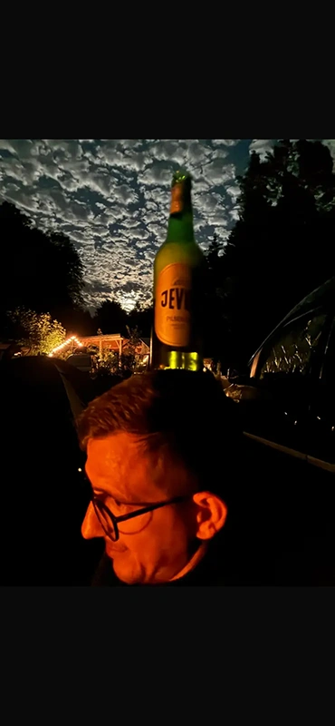 Eine Grüne Bierflasche steht auf einem Kopf. Im Hintergrund sind Bäume, Lichter und ein bewölkter Nachthimmel zu sehen.