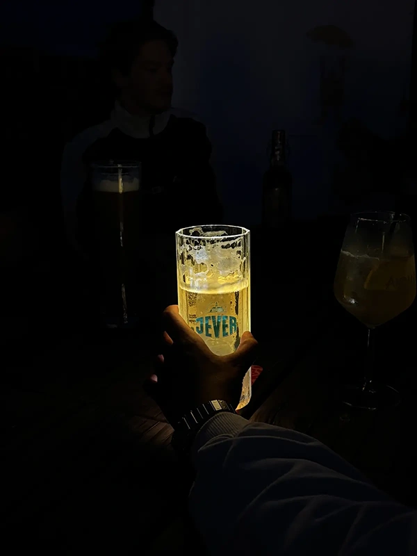 Hand hält ein beleuchtetes Bierglas mit Jever Logo. Im Hintergrund sitzen Personen an einem dunklen Holztisch.