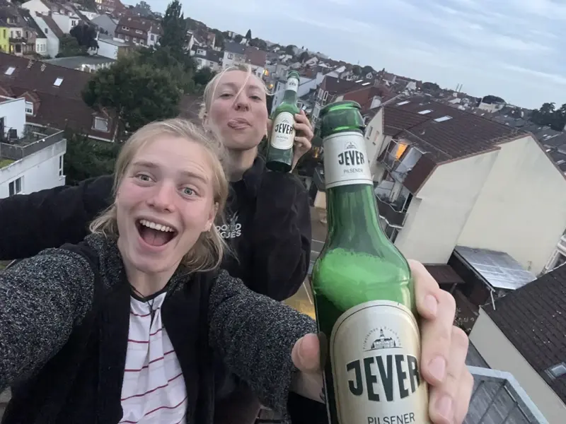 Zwei Personen halten Bierflaschen mit Jever Etikett in die Kamera. Im Hintergrund sind Häuserdächer einer Stadt zu sehen.