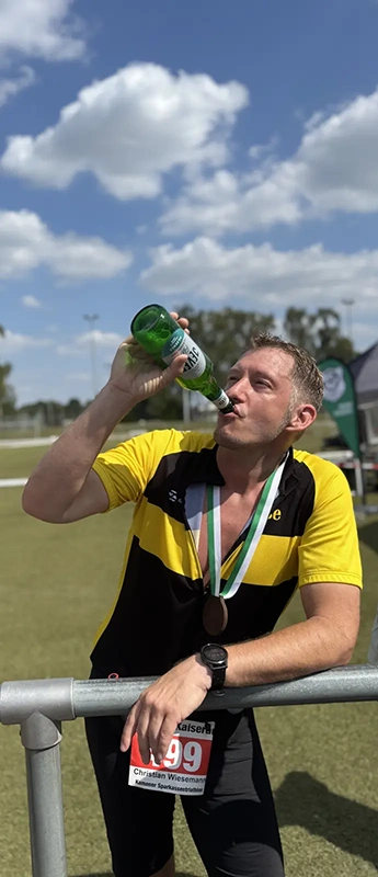 Mann in gelb-schwarzem Sporttrikot trinkt aus einer Bierflasche. Im Hintergrund sind Sportplatz, Bäume und blauer Himmel mit Wolken.