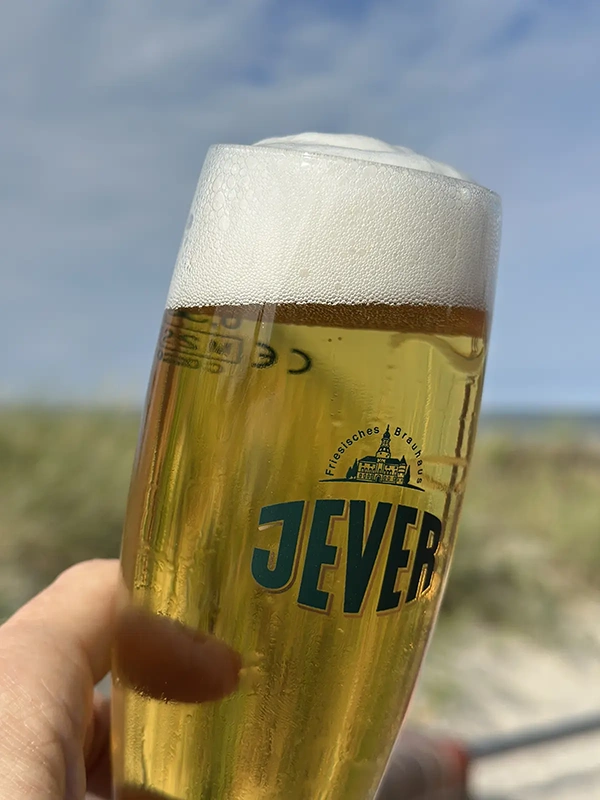 Hand hält ein gefülltes Bierglas mit Jever Logo. Im Hintergrund sind unscharfe Dünen und ein blauer Himmel zu sehen.