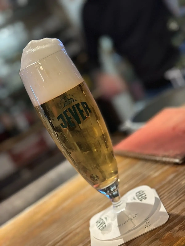 Gefülltes Bierglas mit Jever Logo steht auf einem Bierdeckel. Im unscharfen Hintergrund ist eine Person an einer Theke zu sehen.
