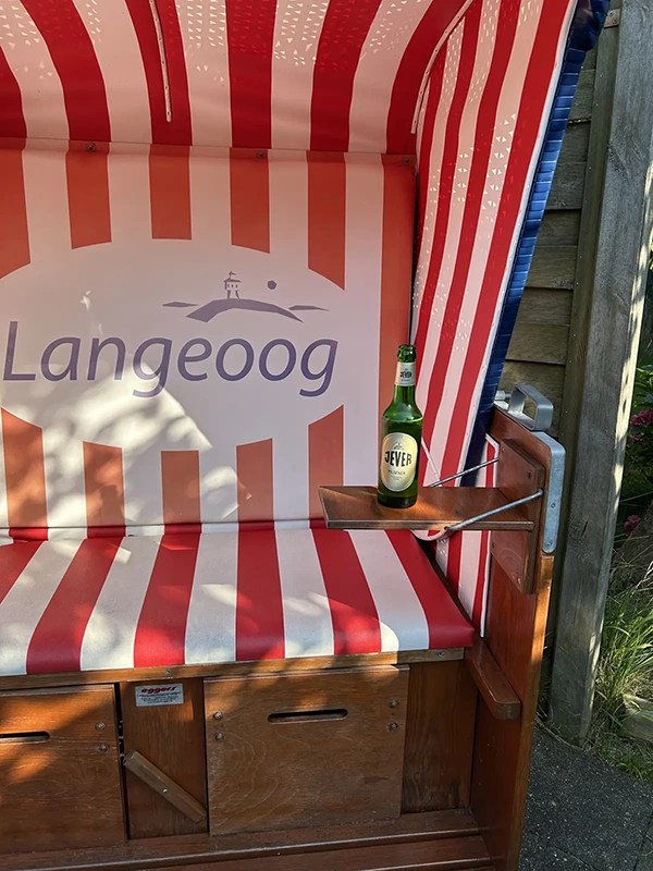 Grüne Bierflasche mit Jever Etikett steht auf einer kleinen Ablage. Dahinter befindet sich ein rot-weiß gestreifter Strandkorb mit Aufschrift Langeoog.
