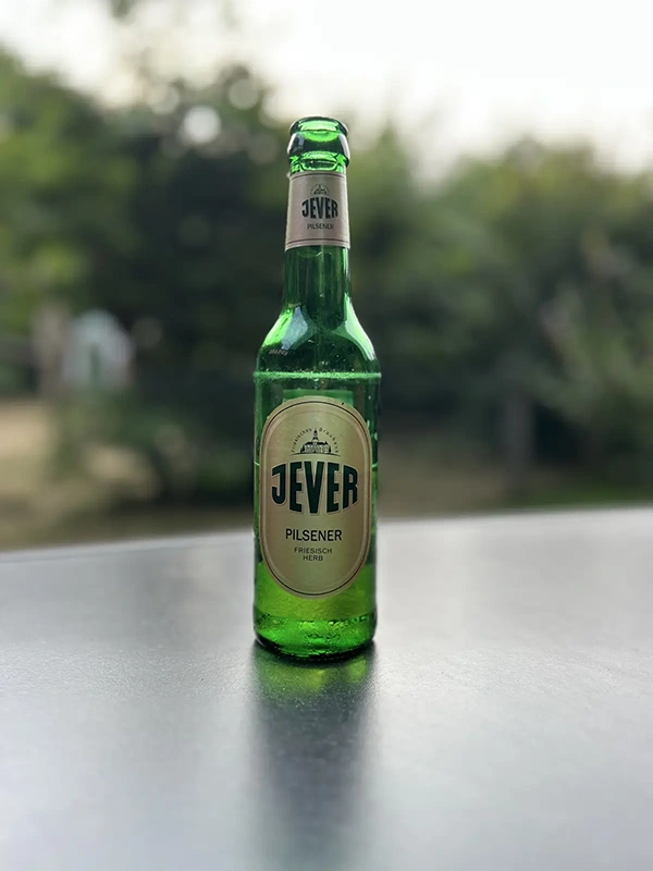 Grüne Bierflasche mit Jever Etikett steht auf einer grauen Tischplatte. Der unscharfe Hintergrund zeigt Bäume und hellen Himmel.