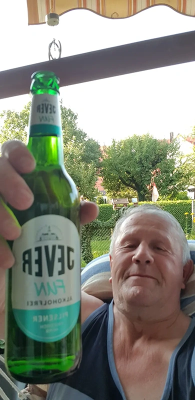 Mann hält eine grüne Bierflasche mit Jever Etikett in die Kamera. Im Hintergrund sind Bäume, Häuser und ein heller Himmel zu sehen.