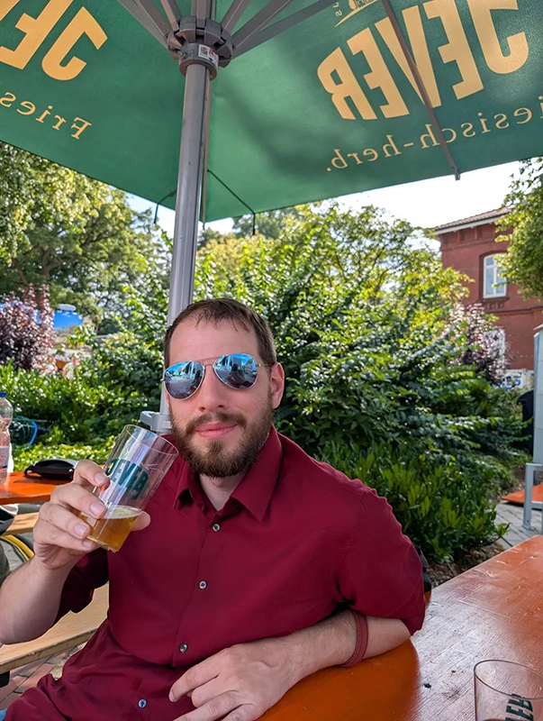 Mann mit Sonnenbrille hält ein Glas mit Jever Logo. Im Hintergrund sind grüne Sträucher, Bäume und ein großer Sonnenschirm zu sehen.
