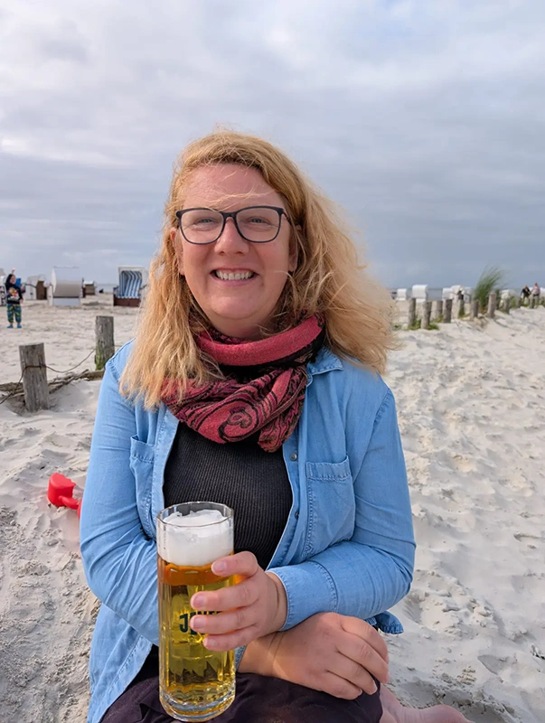 Frau mit Brille hält ein Glas Bier mit Jever Logo. Im Hintergrund sind Sandstrand, Strandkörbe und ein bewölkter Himmel zu sehen.