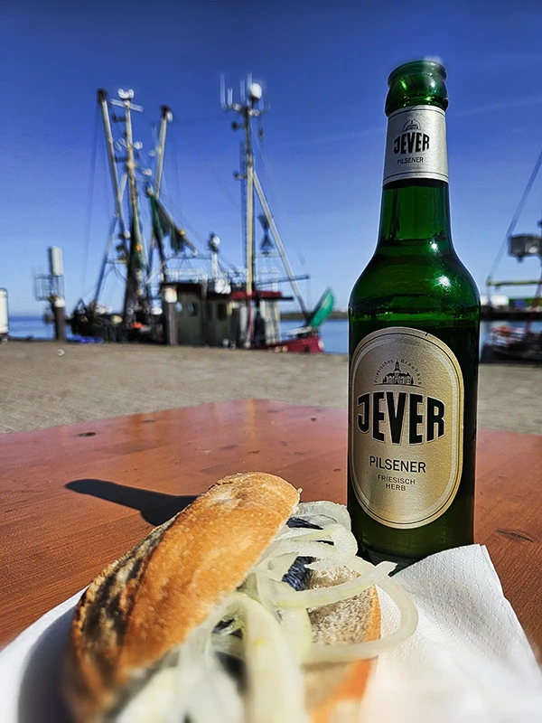 Grüne Bierflasche mit Jever Etikett steht auf einem Holztisch neben einem Fischbrötchen. Im Hintergrund sind Fischerboote und blauer Himmel zu sehen.