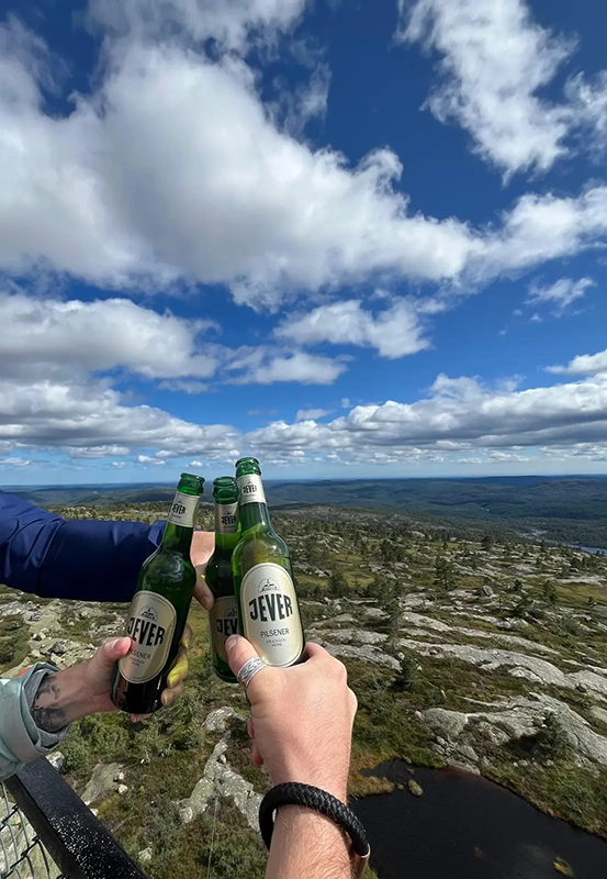 Drei Personen stoßen mit Jever Bierflaschen an. Im Hintergrund sind eine felsige Hügellandschaft und ein teils bewölkter Himmel zu sehen.