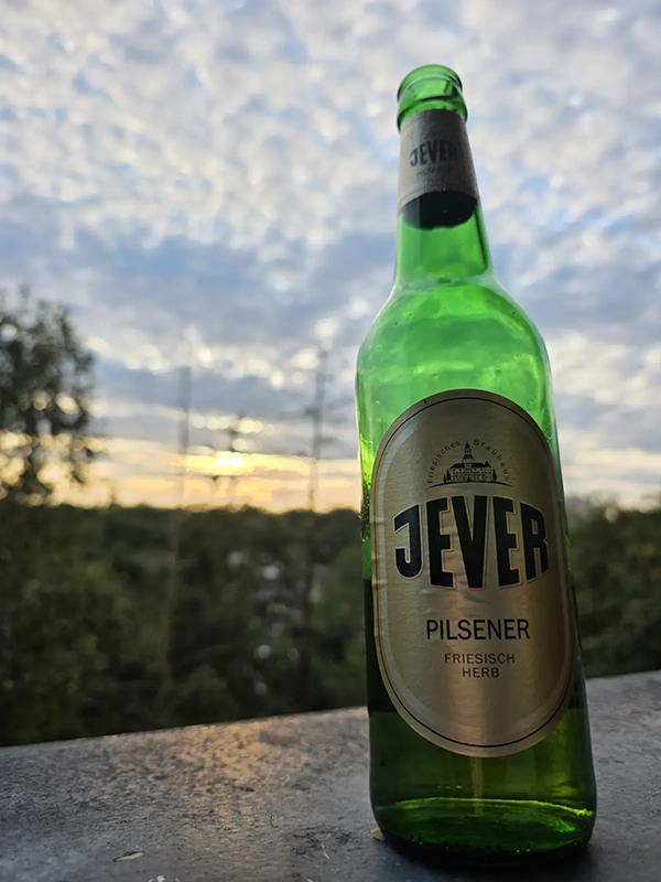 Grüne Bierflasche mit Jever Etikett steht auf einer Steinfläche. Im Hintergrund sind Bäume und ein Himmel mit dichter Wolkendecke bei Sonnenuntergang zu sehen.