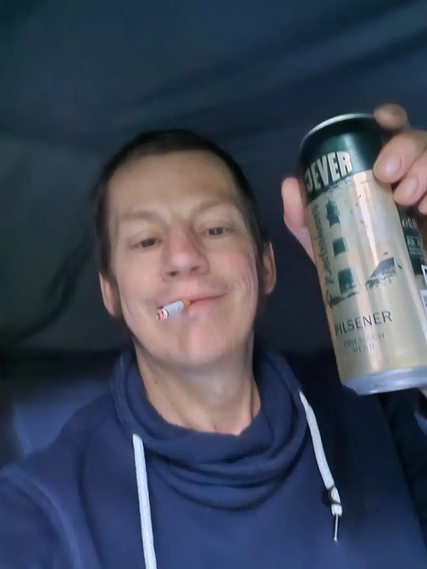 Person im blauen Pullover hält eine Jever Bierdose hoch. Im Mund befindet sich eine brennende Zigarette, der Hintergrund ist unscharf.