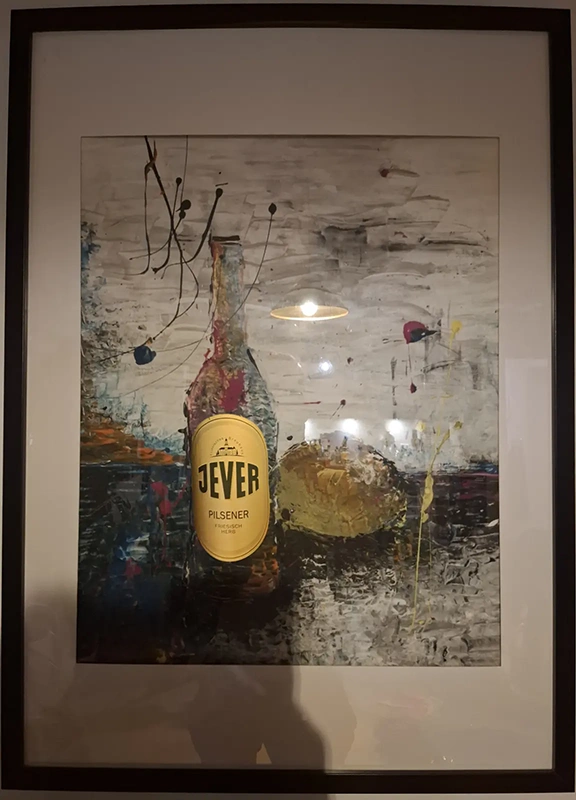 Gerahmtes Bild mit abstrakter Malerei einer Flasche und eines Brotes. Auf der Flasche ist ein echtes Jever Etikett angebracht.