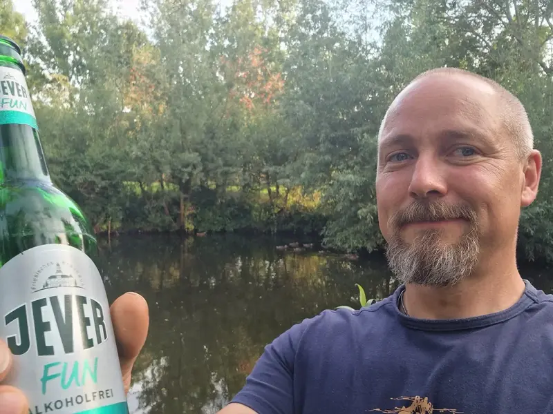 Mann hält eine grüne Bierflasche mit Jever Etikett in die Kamera. Im Hintergrund ist ein Gewässer mit dicht bewachsenen Ufern zu sehen.