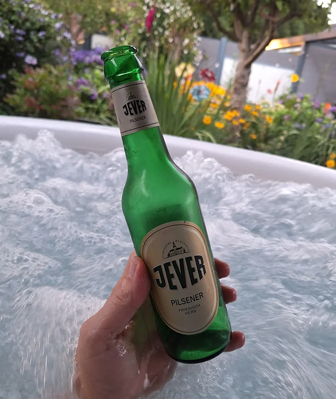 Grüne Bierflasche Jever Pilsener wird in einer Hand über sprudelndem Wasser gehalten. Im Hintergrund blühende Gartenpflanzen.