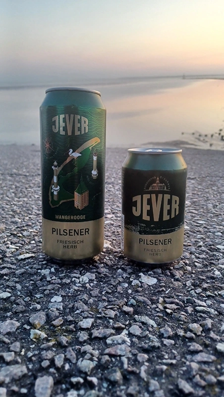 Zwei Jever Bierdosen stehen auf Asphalt am Ufer. Im Hintergrund ein Sonnenuntergang über dem Wasser.