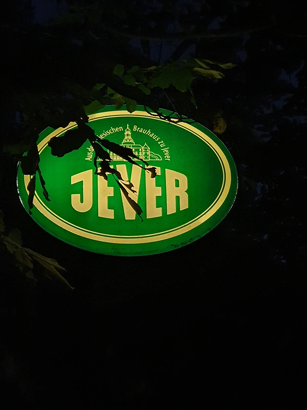 Ein grünes Jever Leuchtschild hängt in der Dunkelheit. Im Vordergrund sind Blätter von Bäumen zu erkennen.