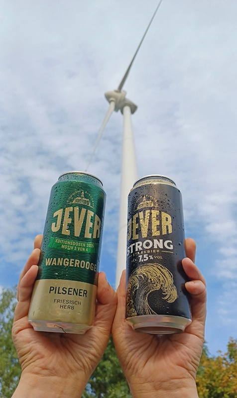 Zwei Jever Dosen werden in den Händen hochgehalten. Im Hintergrund ein Windrad und blauer Himmel.