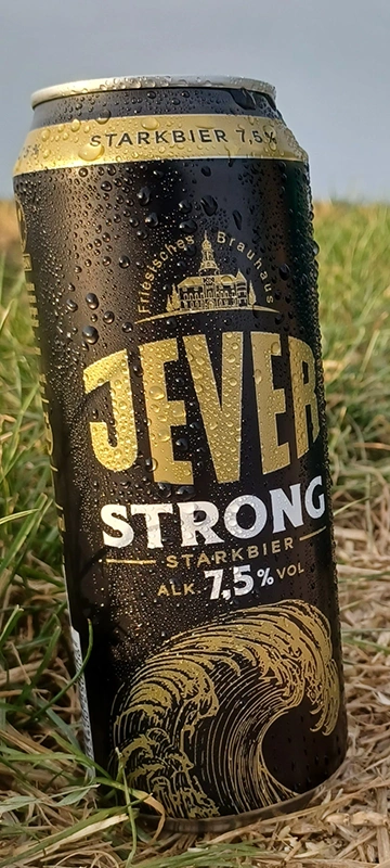 Eine schwarze Jever Strong Dose mit Wassertropfen steht auf Gras. Dahinter unscharf der Himmel.