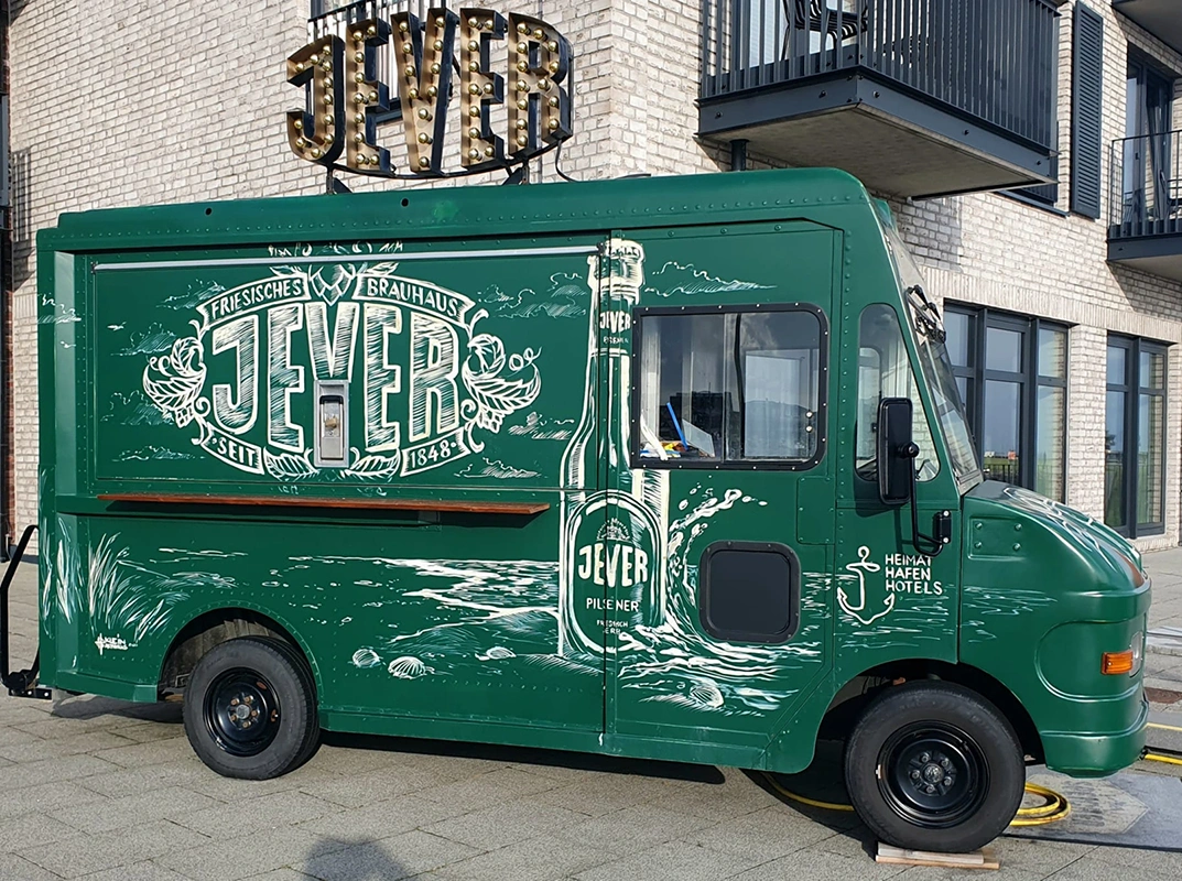 Ein grüner Foodtruck mit großem Jever Schriftzug und großer illustrierter Bierflasche steht vor einem Gebäude.