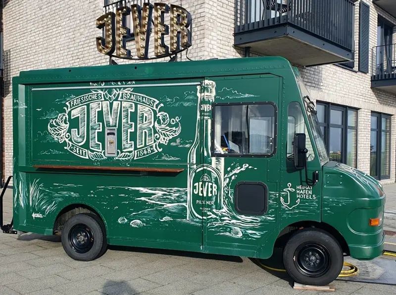 Ein grüner Foodtruck mit großem Jever Schriftzug und großer illustrierter Bierflasche steht vor einem Gebäude.