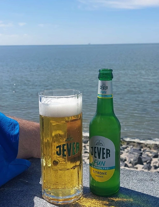 Ein Glas mit Jever Bier und eine Jever Fun Zitrone Alkoholfrei Flasche stehen auf einer Mauer. Dahinter das Meer.
