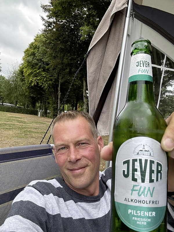 Ein Mann hält eine Jever Fun Alkoholfrei Flasche in die Kamera. Im Hintergrund Bäume und ein Zelt.