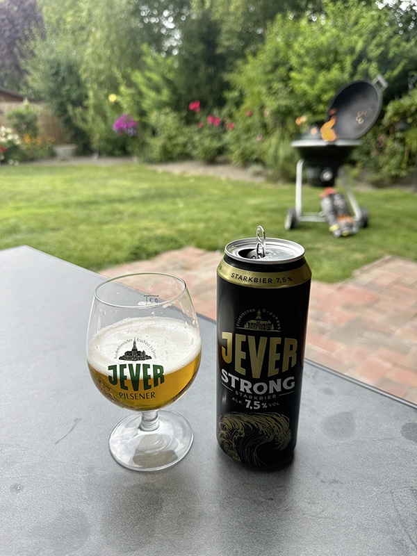 Ein Glas mit Jever Bier und eine geöffnete Jever Strong Dose stehen auf einem Tisch. Im Hintergrund ein Grill im Garten.