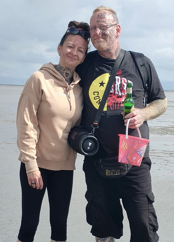 Ein Paar steht am Strand, der Mann hält eine Jever Bierflasche und einen bunten Eimer. Im Hintergrund ist das Watt erkennbar.