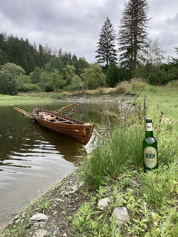 Eine Jever Bierflasche steht im Gras am Ufer neben einem Holzboot im Wasser. Im Hintergrund sind Bäume und ein Hochsitz.