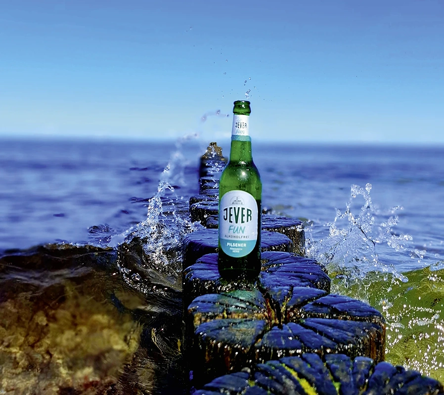 Eine Jever Bierflasche steht auf nassen Holzpfählen am Meer. Im Hintergrund spritzt Wasser und der Himmel ist blau.