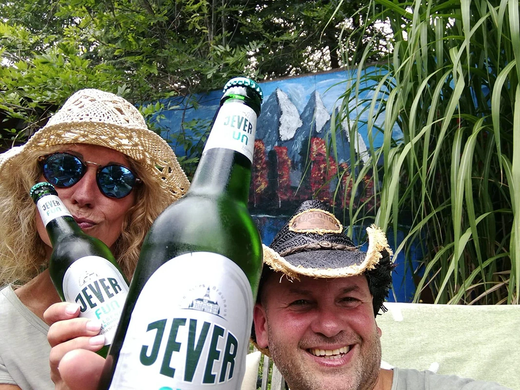 Zwei Personen mit Strohhüten halten Jever Bierflaschen in die Kamera. Im Hintergrund Pflanzen und eine bemalte Wand.