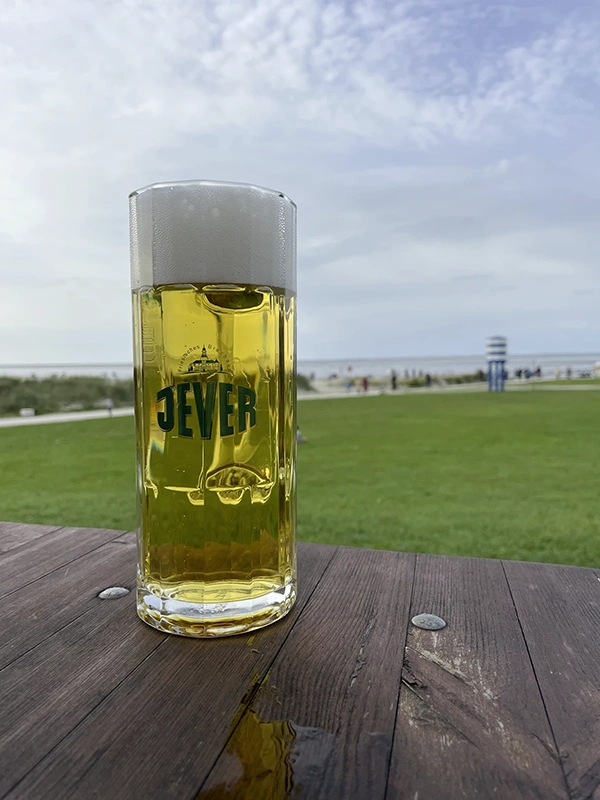 Ein gefülltes Bierglas steht auf einem Holztisch. Im Hintergrund eine Wiese mit Strand und Meer.