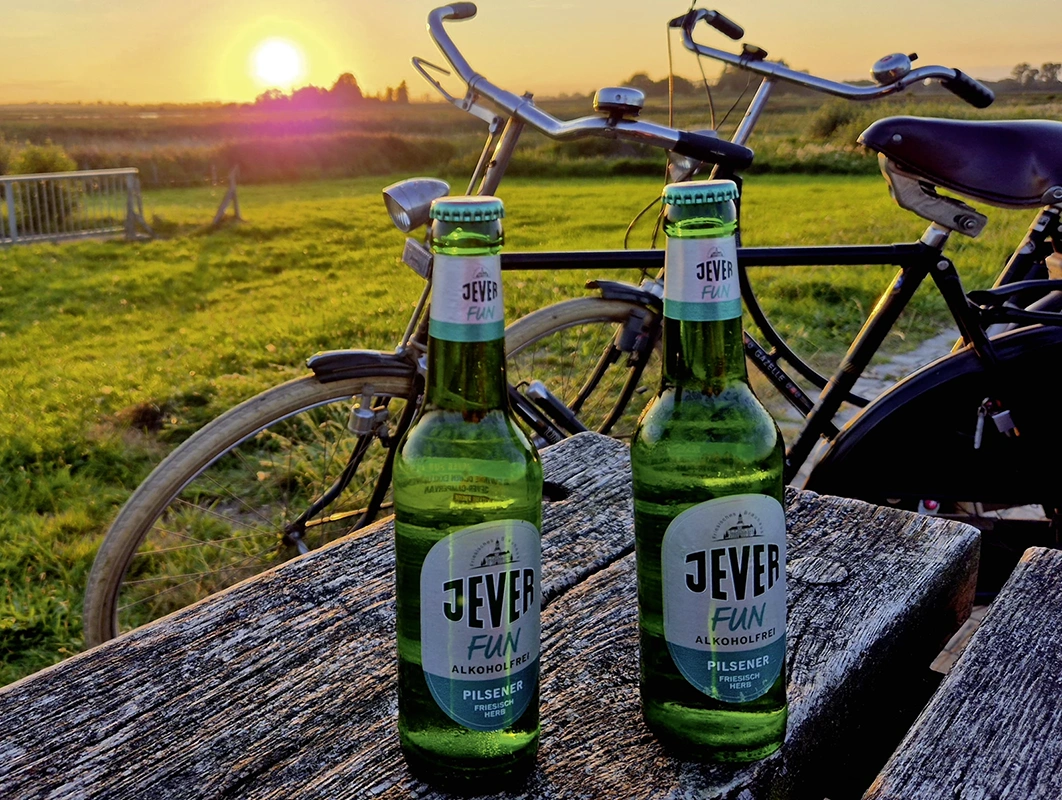 Zwei Jever Bierflaschen stehen auf einem Holztisch. Im Hintergrund sind Fahrräder und ein Sonnenuntergang auf einer Wiese. 