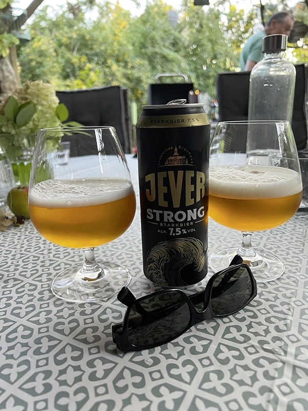 Eine Jever Strong Bierdose steht auf einem Tisch zwischen zwei Gläsern in einem Garten. Davor liegt eine Sonnenbrille.