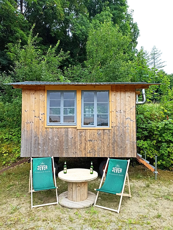 Vor einer Holzhütte stehen zwei grüne Liegestühle mit Jever Aufschrift. Daneben ein runder Holztisch mit zwei Jever Bierflaschen.