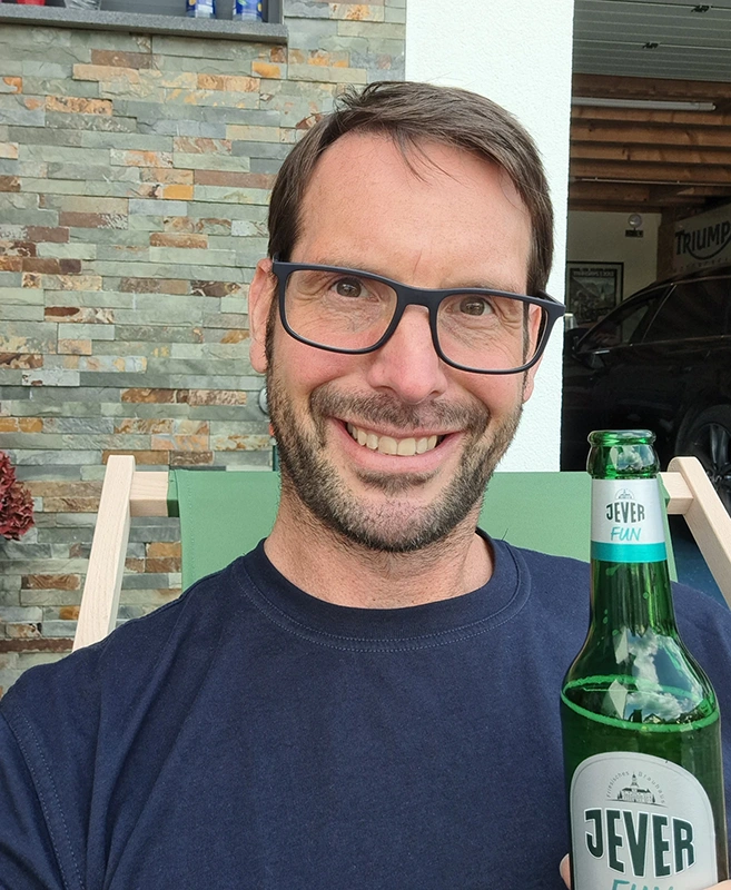 Ein Mann mit Brille hält eine grüne Jever Flasche in der Hand. Hinter ihm eine Steinwand und ein schwarzes Auto.