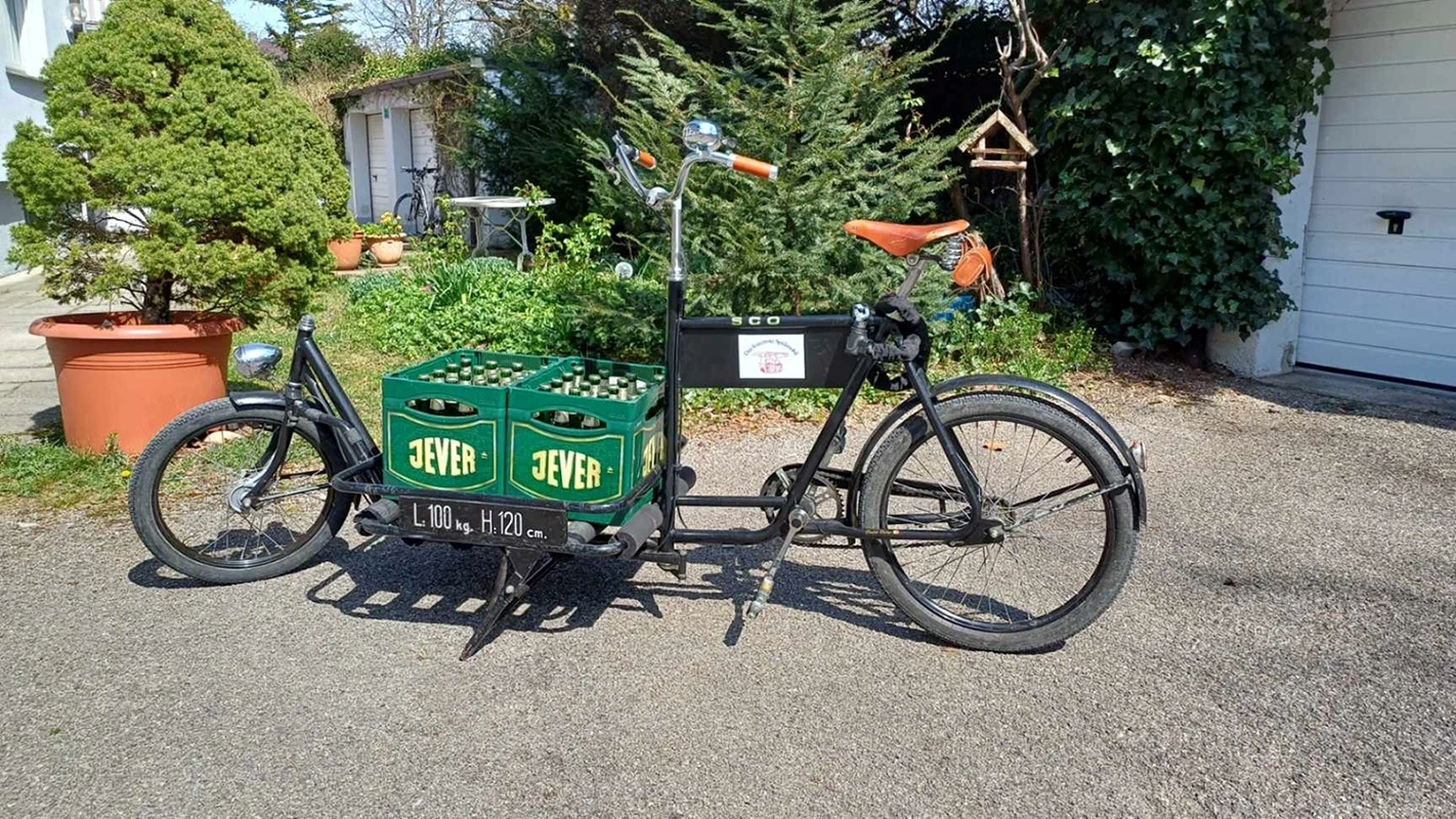 Ein schwarzes Lastenrad transportiert zwei grüne Getränkekisten von Jever. Im Hintergrund sind Bäume, Haus und Garagen.