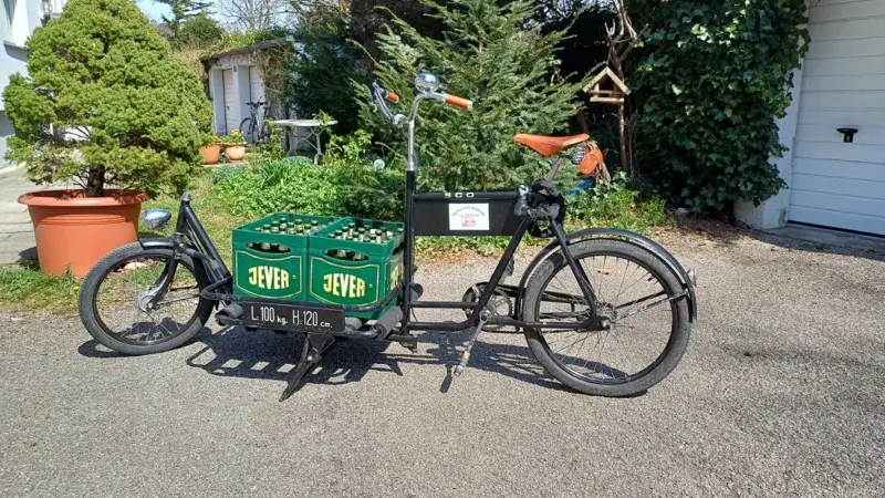 Ein schwarzes Lastenrad transportiert zwei grüne Getränkekisten von Jever. Im Hintergrund sind Bäume, Haus und Garagen.