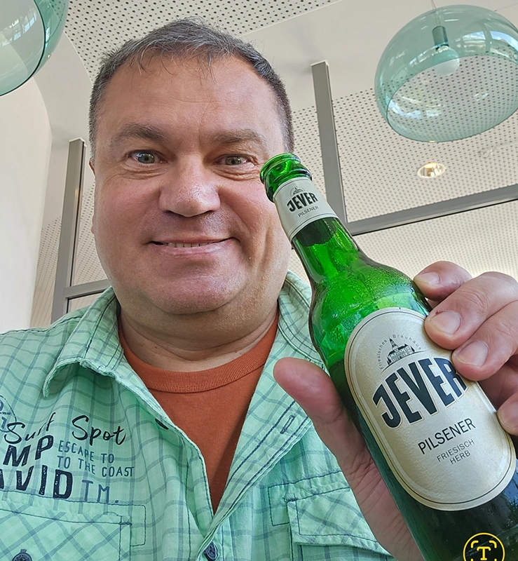 Ein Mann im grünen Hemd hält eine Jever Flasche in der Hand. Hinter ihm sind Lampen und eine helle Decke.