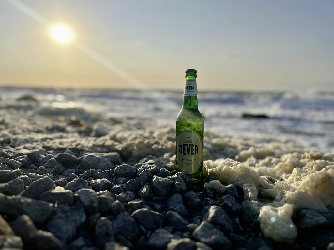 Eine grüne Jever Flasche steht auf Steinen am Strand. Dahinter ist das Meer mit Sonnenuntergang.