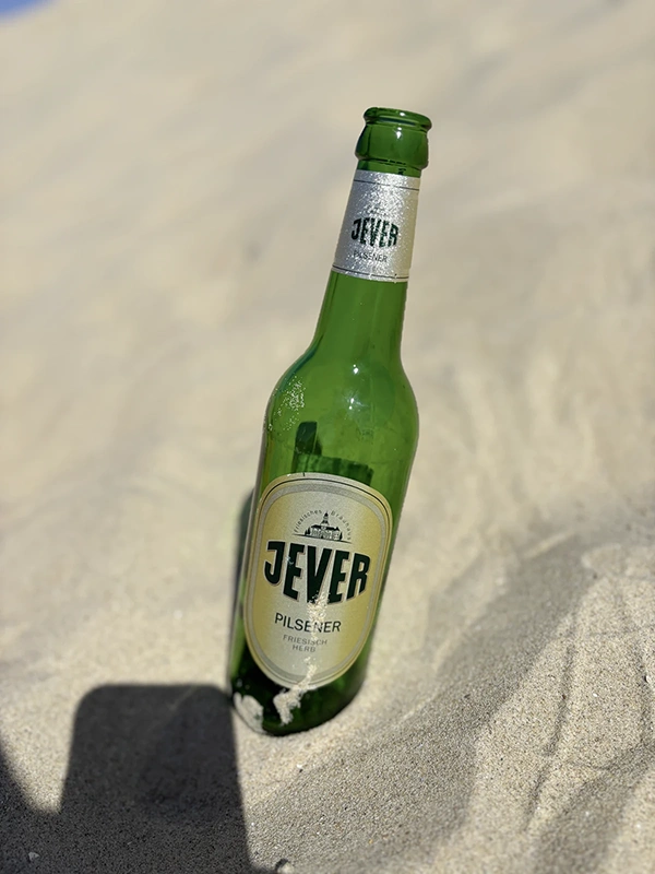 Eine grüne Jever Flasche steckt im hellen Sand. Der Hintergrund ist unscharf und sandig.