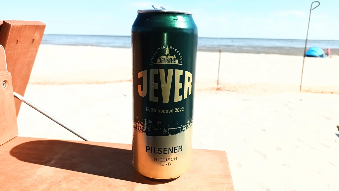 Eine Jever Dose steht auf einem Holztisch. Dahinter liegt ein heller Sandstrand mit Blick auf das Meer.