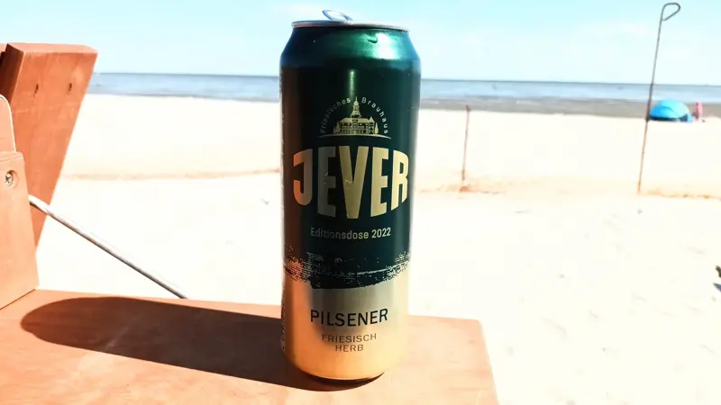 Eine Jever Dose steht auf einem Holztisch. Dahinter liegt ein heller Sandstrand mit Blick auf das Meer.