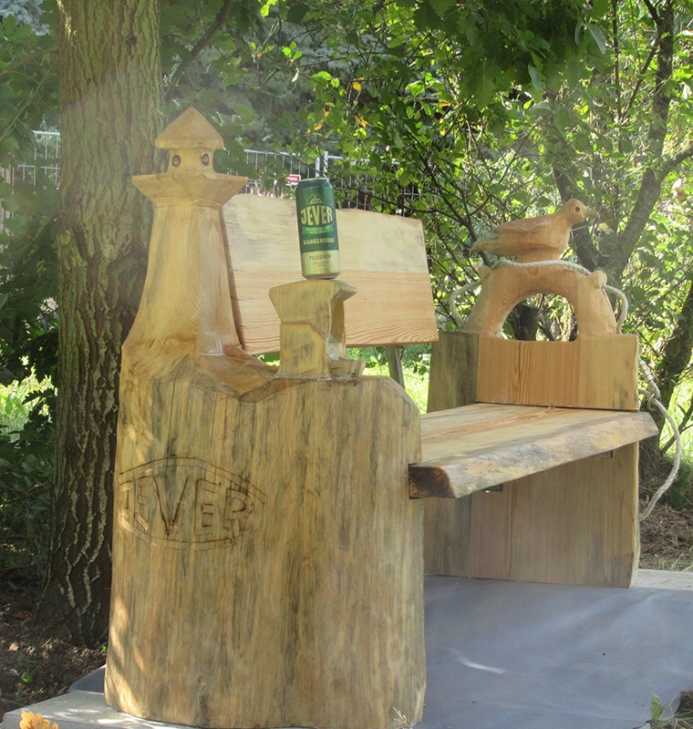 Eine Jever Dose steht auf einer geschnitzten Holzbank mit Jever Prägung und Vogelverzierung. Im Hintergrund Bäume.