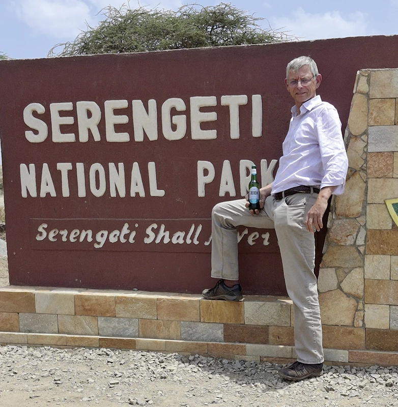 Ein Mann lehnt an einem Schild mit der Aufschrift Serengeti National Park. In der Hand hält er eine Jever Flasche.