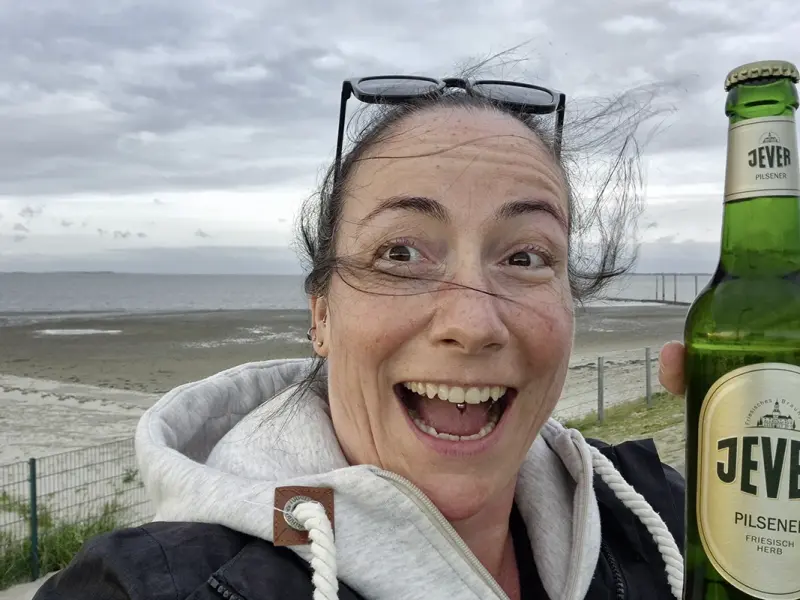 Eine Frau hält strahlend eine Jever Flasche am Strand. Im Hintergrund Meer, Sand und ein bewölkter Himmel.