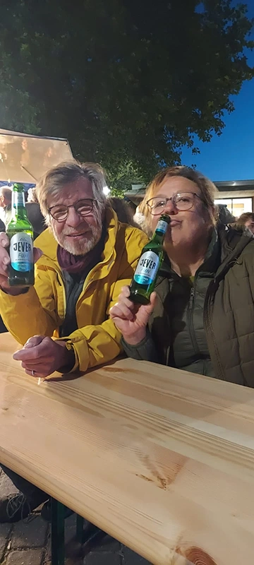 Zwei Personen sitzen an einem Holztisch und halten Jever Flaschen. Im Hintergrund sind weitere Menschen.